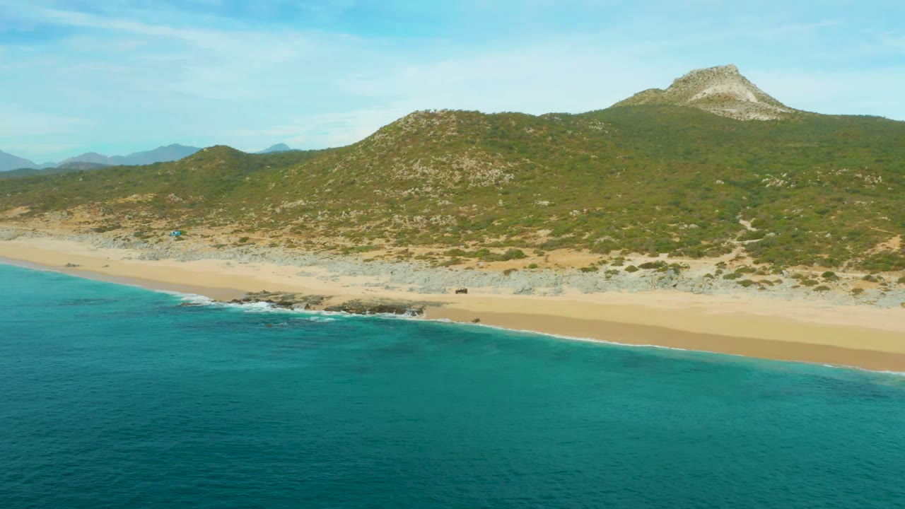 Aerial view at Laguna Beach Los Cabos with turquoise water in the foreground, a pale sandy shore, green rocky hills behind it, and no visible people.