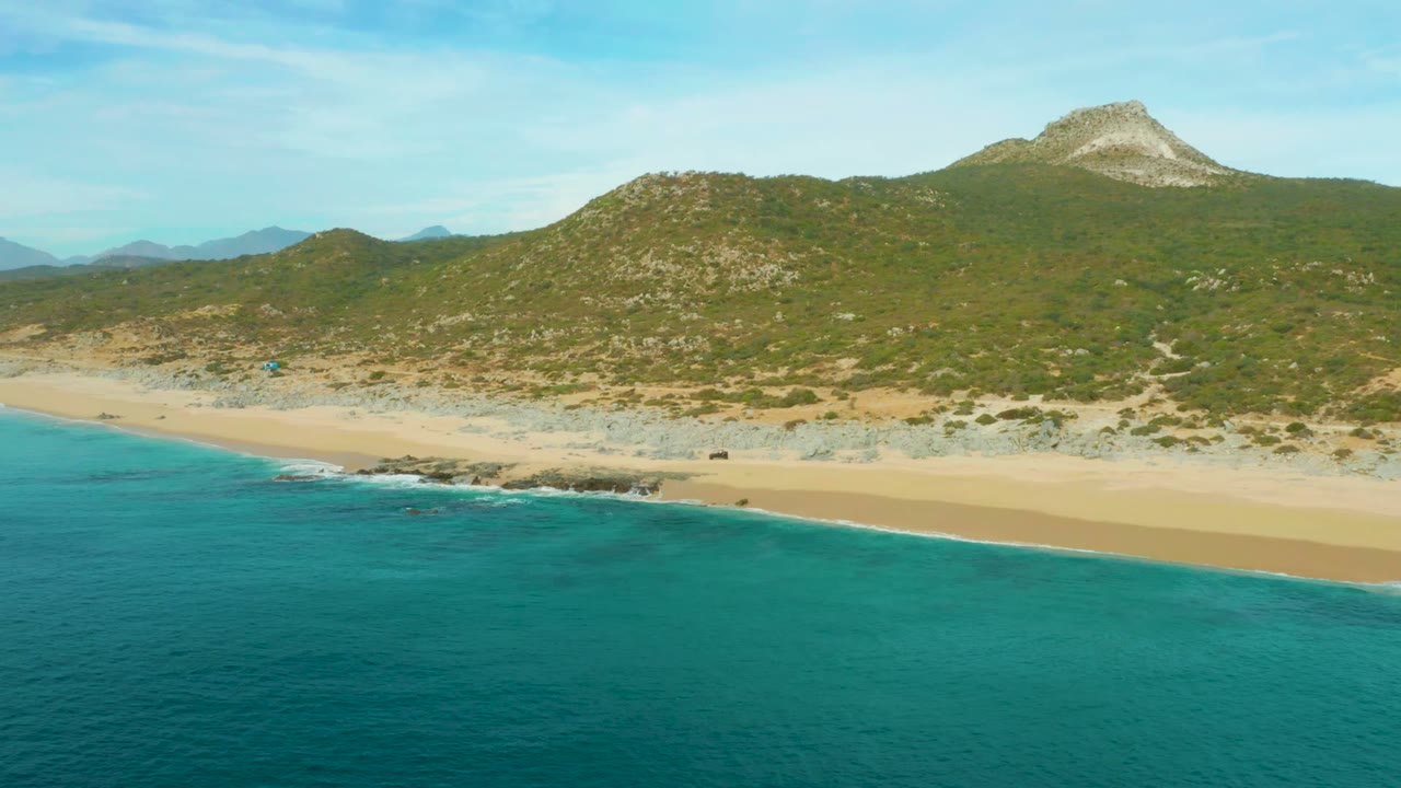 Wide coastal aerial with waves reaching a sandy beach below green hills.