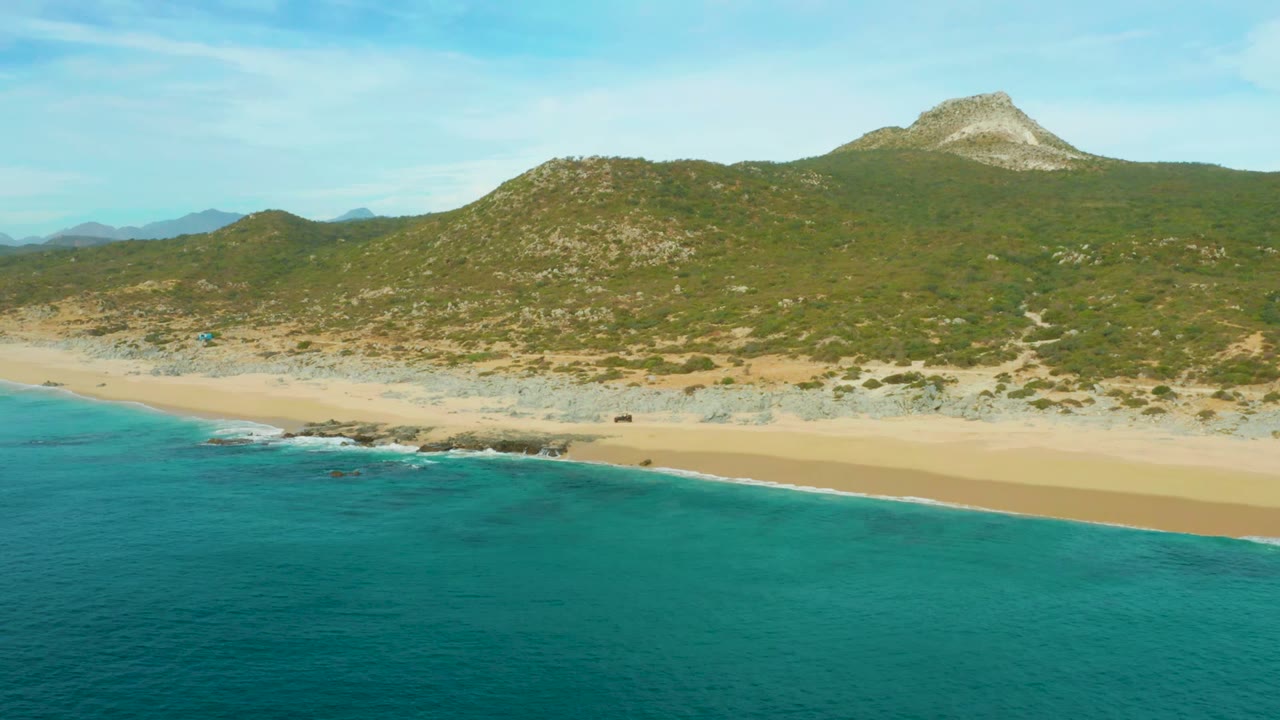 Aerial beach view in Laguna Beach Los Cabos with turquoise sea, a broad empty sand strip, scrub-covered rocky hills, and no visible people.
