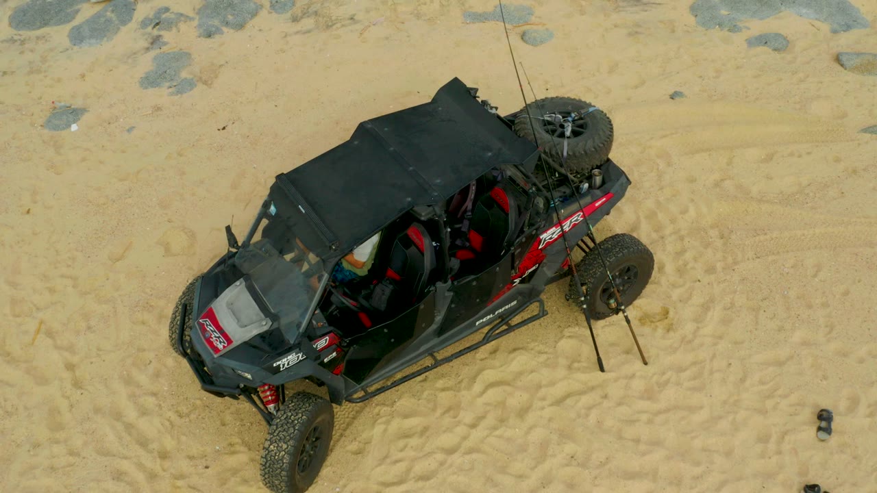 Aerial view of the dune buggy angled across pale sand in Laguna Beach Los Cabos, Mexico, with red interior details, rock patches, and no clearly visible people.