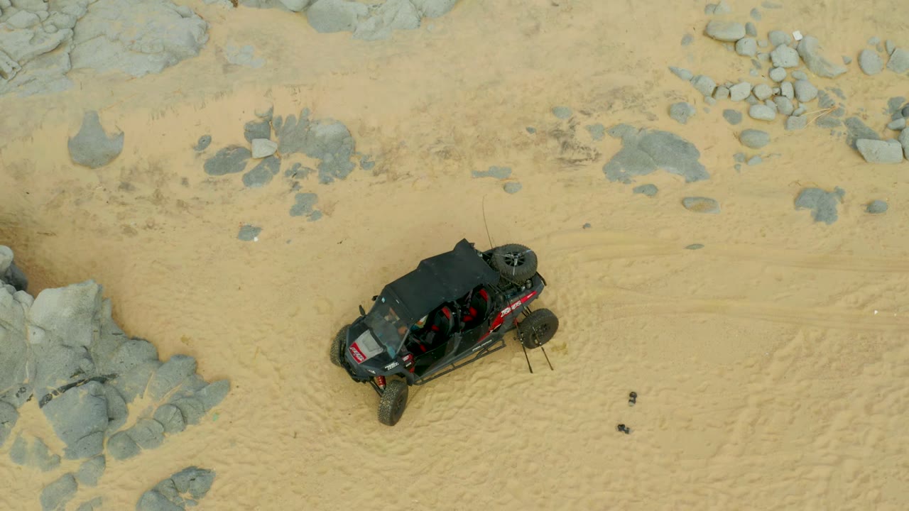 Overhead view of a black off-road buggy on pale sand near gray rock clusters in Laguna Beach Los Cabos, Mexico, with no visible people.