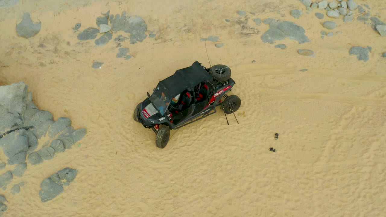 The overhead frame shows the buggy on light sand below scattered gray rocks in Laguna Beach Los Cabos, Mexico, with no visible people.