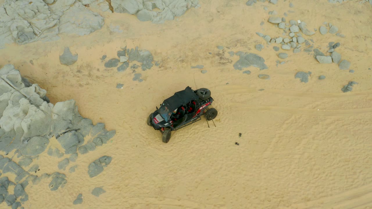 A high overhead view in Laguna Beach Los Cabos shows a black off-road vehicle near gray rocks on pale sand, with tire marks and no visible people.