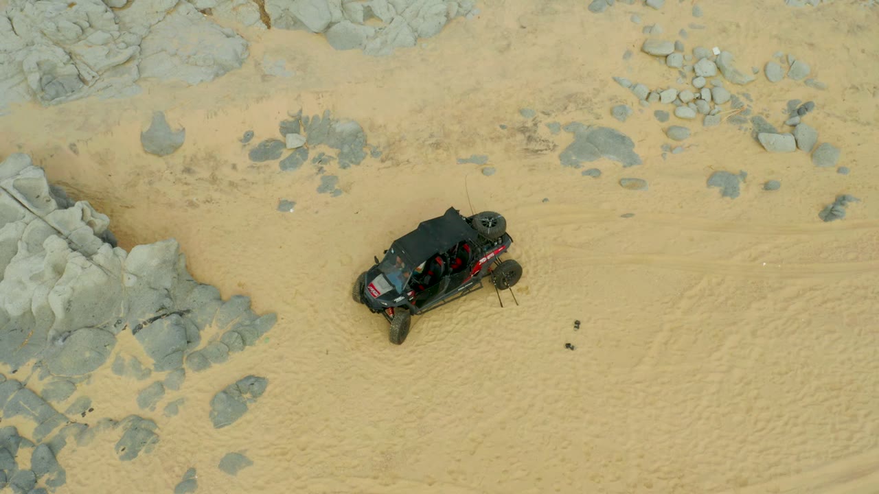 Aerial footage shows the black buggy on pale sand below gray rock patches in Laguna Beach Los Cabos, with the vehicle angled across the open ground and no visible people.