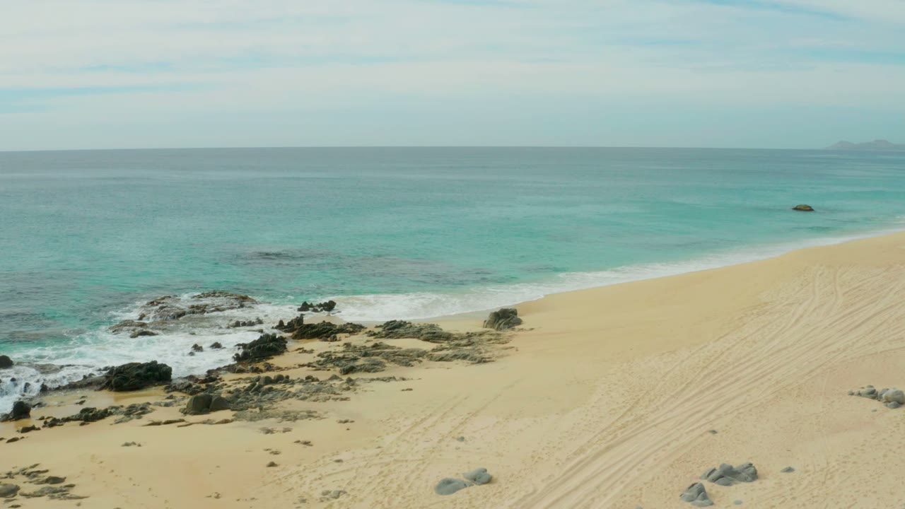 Laguna Beach Los Cabos shoreline with pale sand in the foreground, dark rocks at the waterline, turquoise ocean beyond, and no visible people.