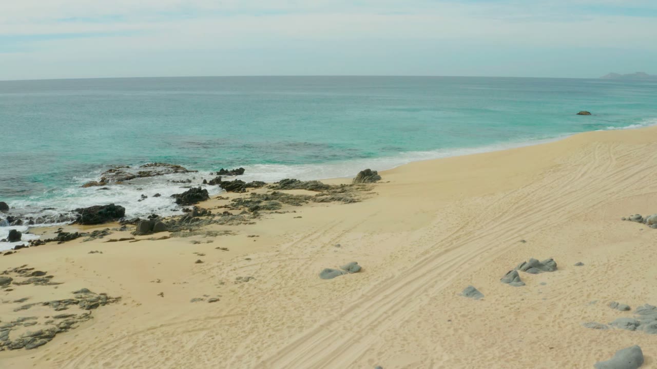 Laguna Beach Los Cabos sand with diagonal tire tracks, rounded boulders in the foreground, rocky surf in the midground, turquoise water, and no visible people.