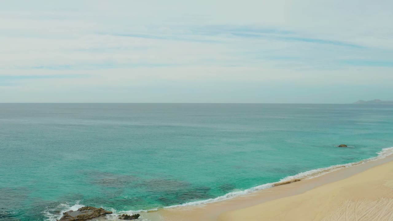 Elevated view of Laguna Beach Los Cabos with turquoise water, pale sand, dark rocks at the lower left, a level horizon, and no visible people.