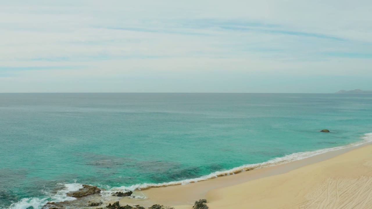 Small waves breaking around rocks beside a wide sandy beach.