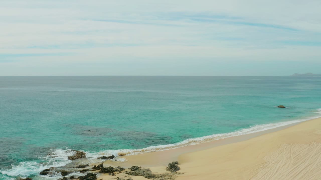 Elevated Laguna Beach Los Cabos view with foamy surf over shoreline rocks, a long pale sand strip, open turquoise sea, and no visible people.