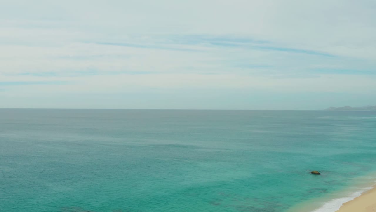 Opening frame of Laguna Beach Los Cabos with turquoise water across the foreground, a small sandy shoreline at lower right, distant land on the horizon, and no visible people.