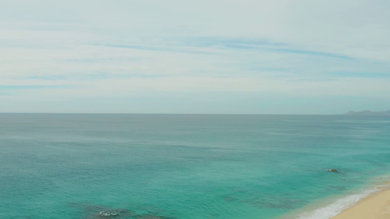 A wider-looking frame from Laguna Beach Los Cabos shows shallow turquoise water near the lower right beach edge, a calm horizon, distant land, and no visible people.