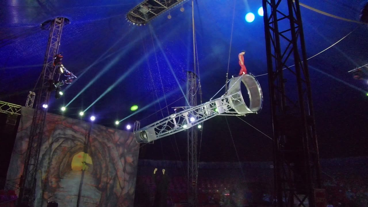 In San Jose del Cabo, a performer stands on a large aerial hoop angled over a circus stage while blue and green lights cross the tent roof.
