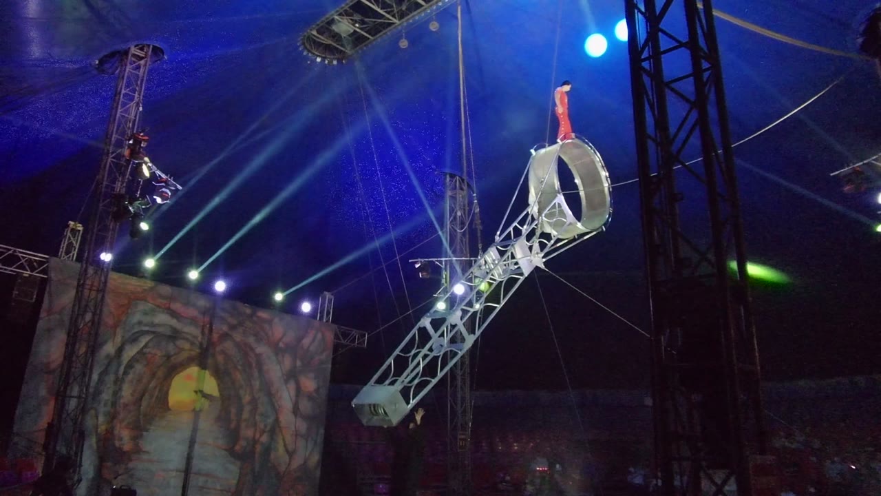 Aerial hoop rotating upward with performer standing on top in a circus tent.