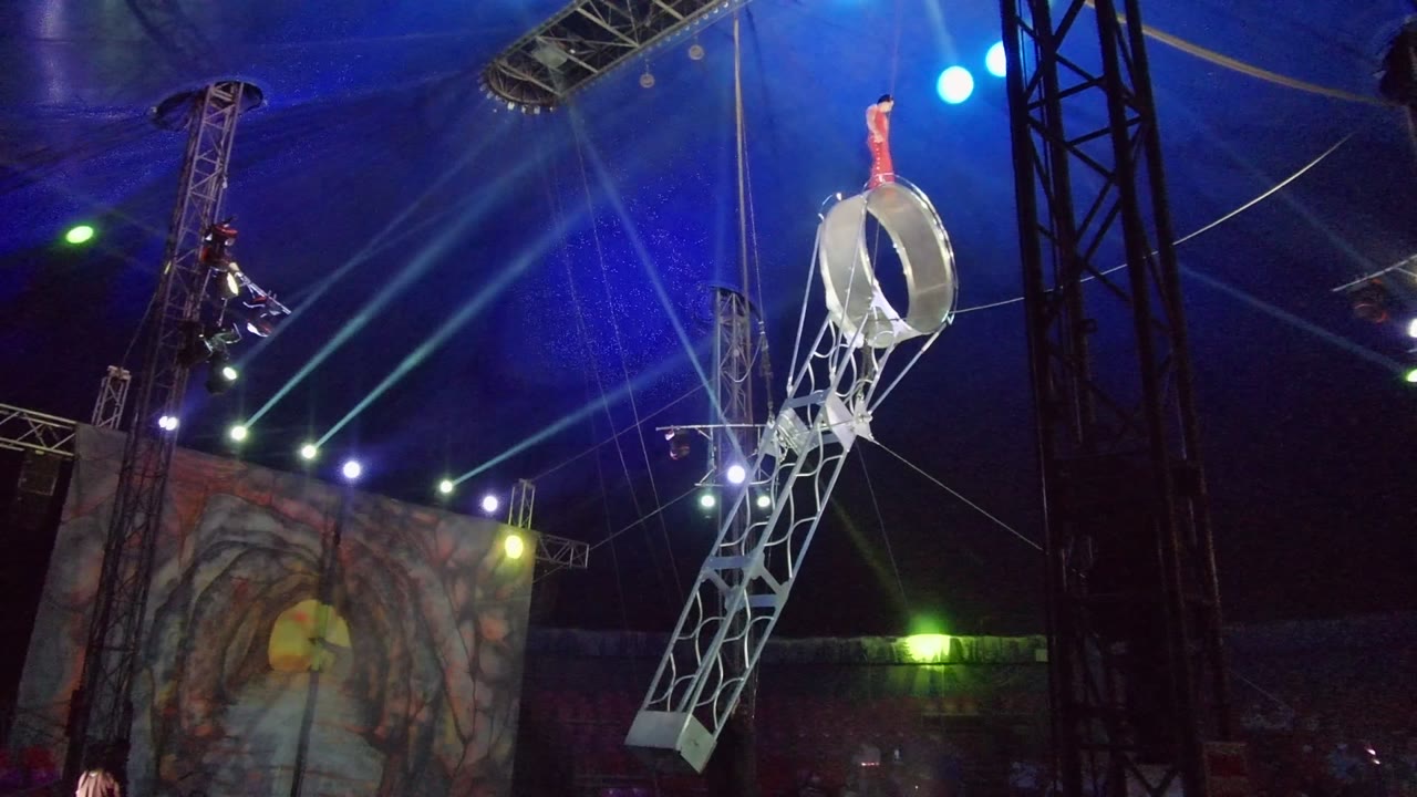 A performer in red stands on the top edge of the nearly upright aerial hoop, with stage trusses, blue lamps, and a cave-like painted backdrop below in San Jose del Cabo.