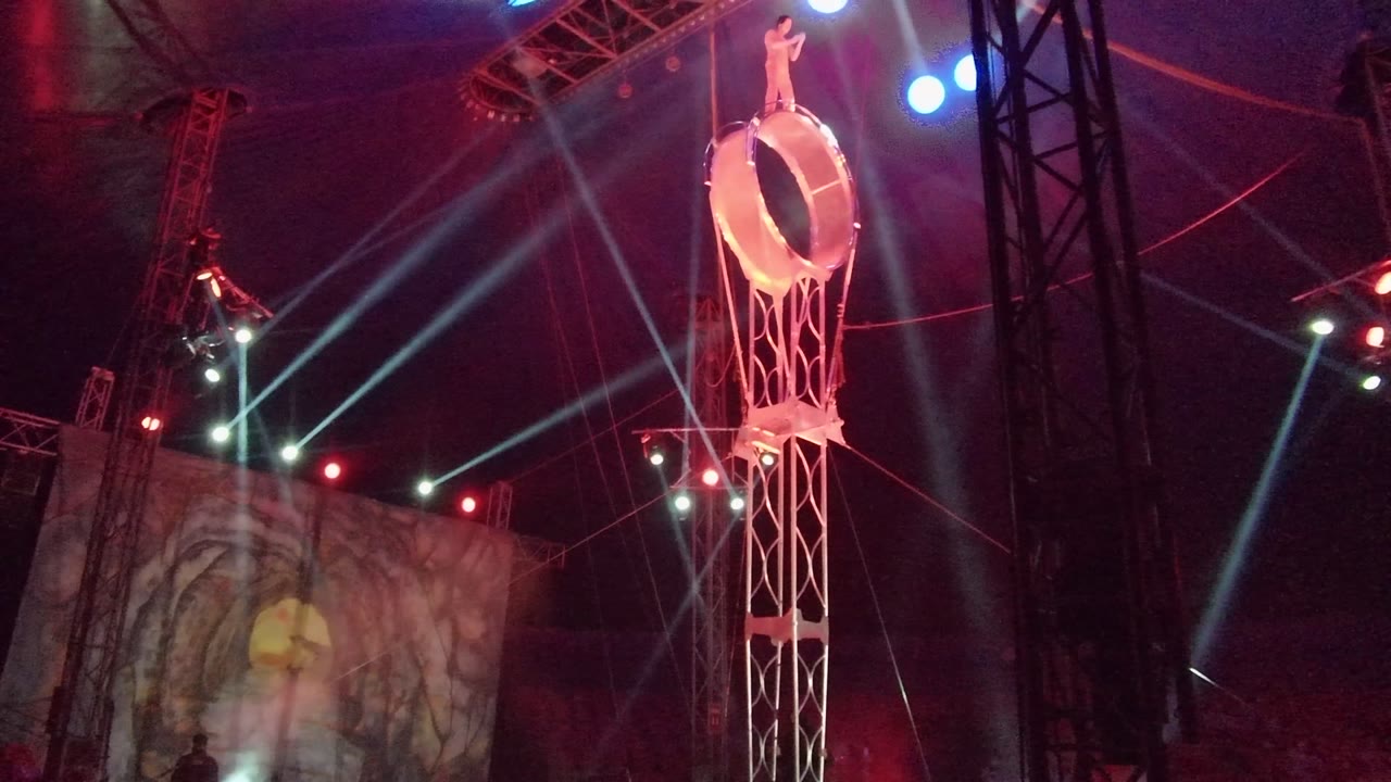 In San Jose del Cabo, a performer stands on top of a tall circular ring structure while red stage light and blue lamps illuminate truss and rigging around the stage.