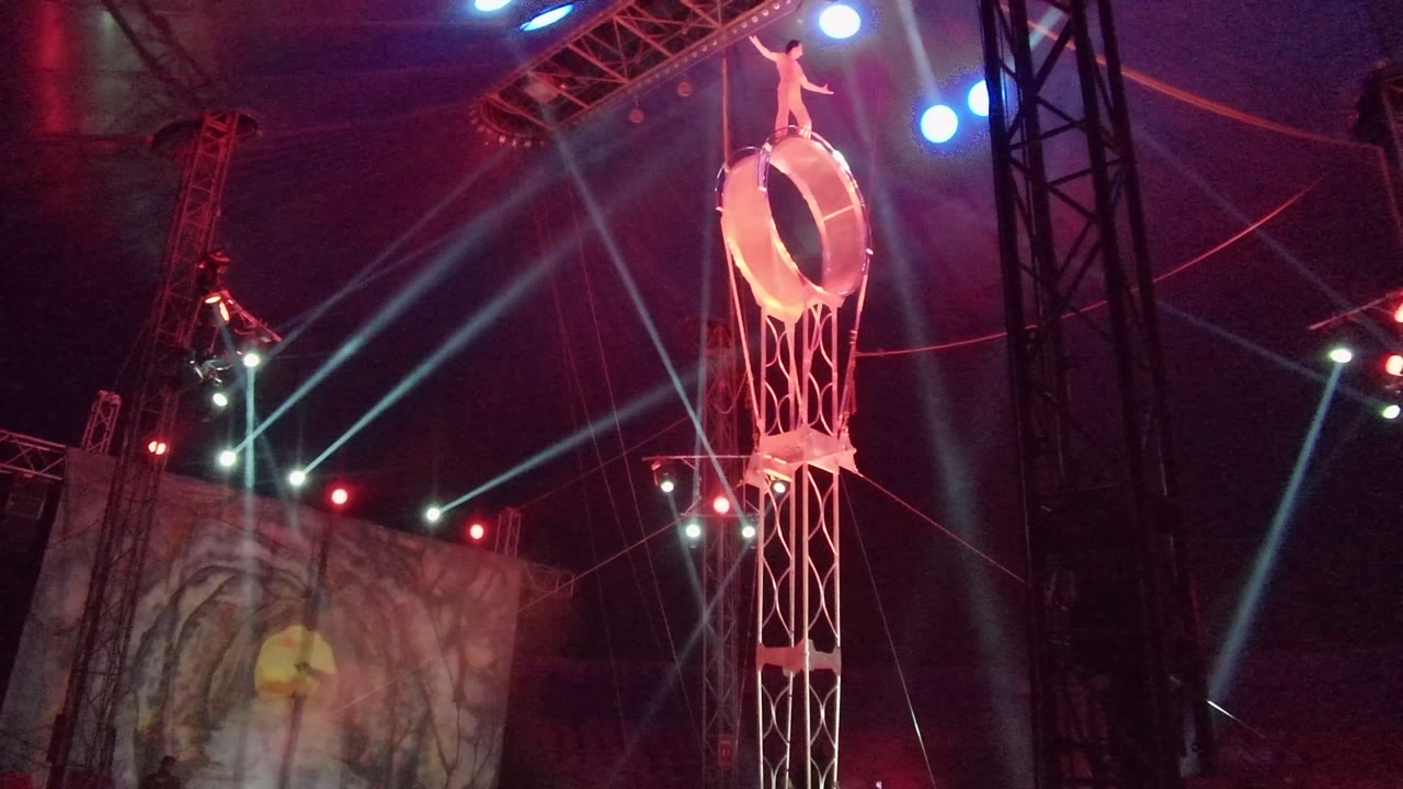 A performer stands at the top of the circular aerial ring in San Jose del Cabo, with the tower near the right side of frame and a dim stage projection visible to the left.