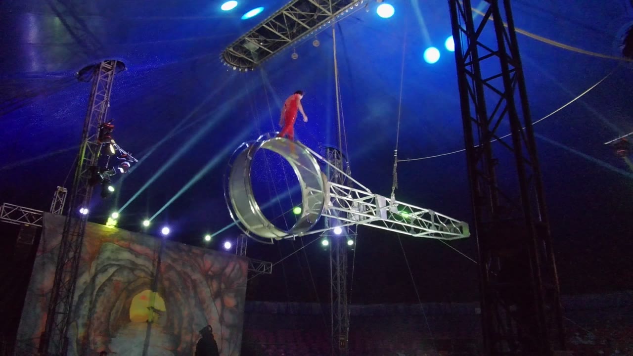 In San Jose Del Cabo, Mexico, a performer in red stands on the upper structure of a suspended aerial ring while blue tent lighting and rigging fill the background.