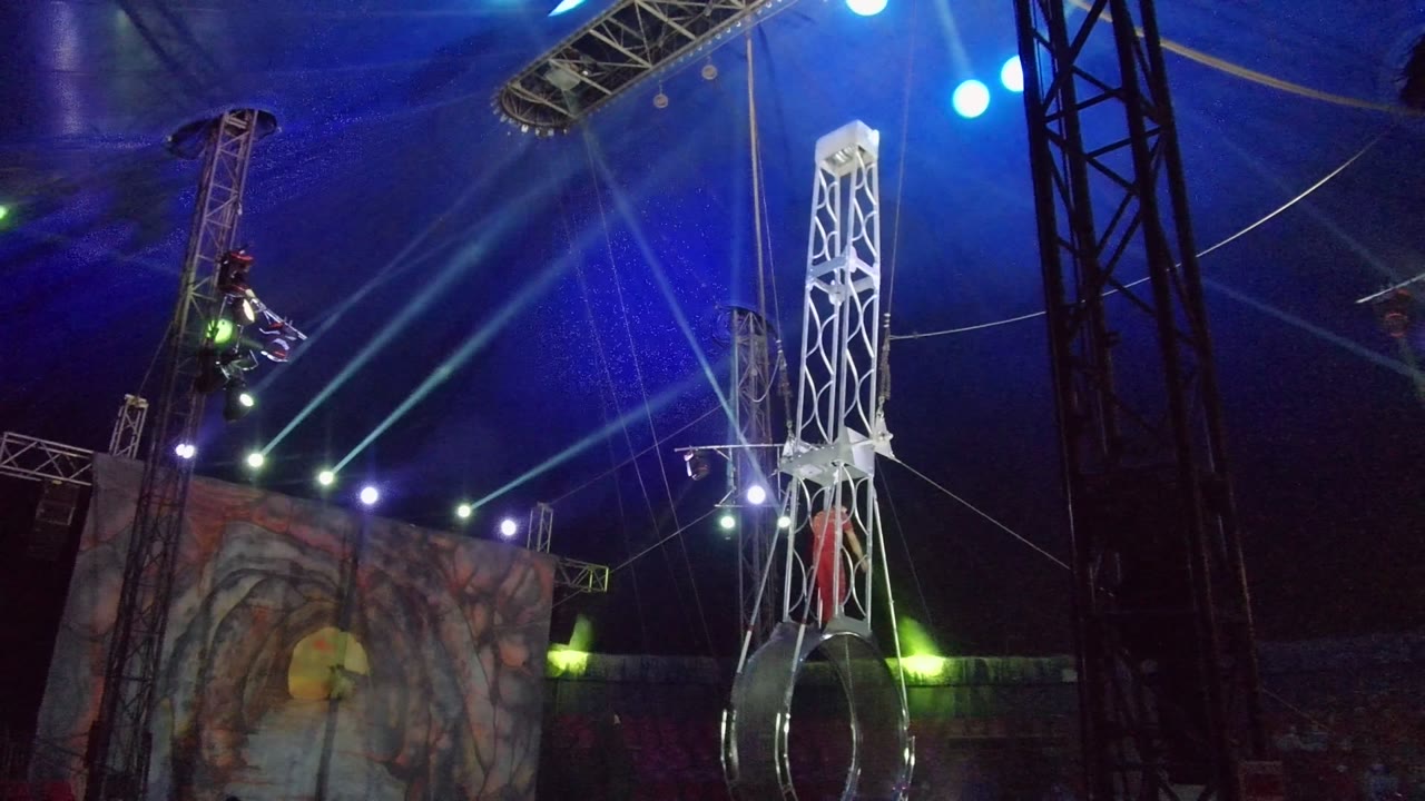 In San Jose Del Cabo, Mexico, the aerial ring hangs below a near-vertical white truss as the performer in red holds position beside the apparatus under colored stage lighting.