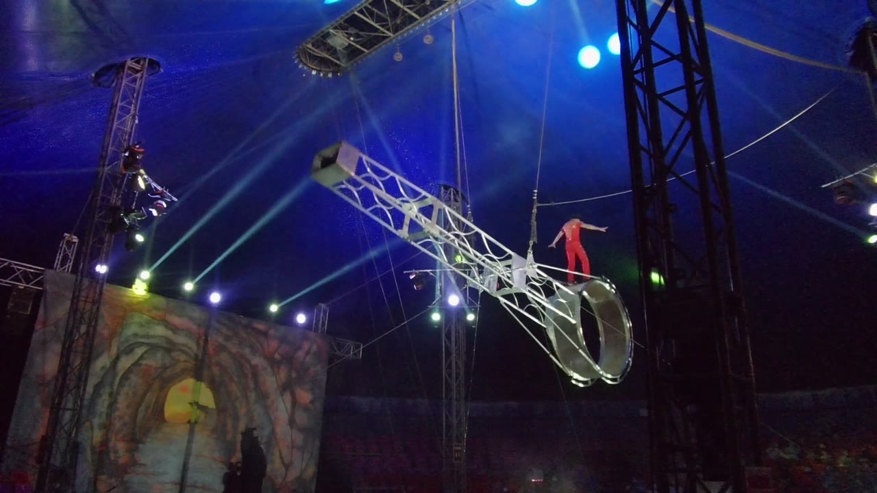 In San Jose del Cabo, an acrobat in red stands on a tilted silver circular apparatus while blue stage lighting, rigging, and a painted backdrop fill the tent interior.