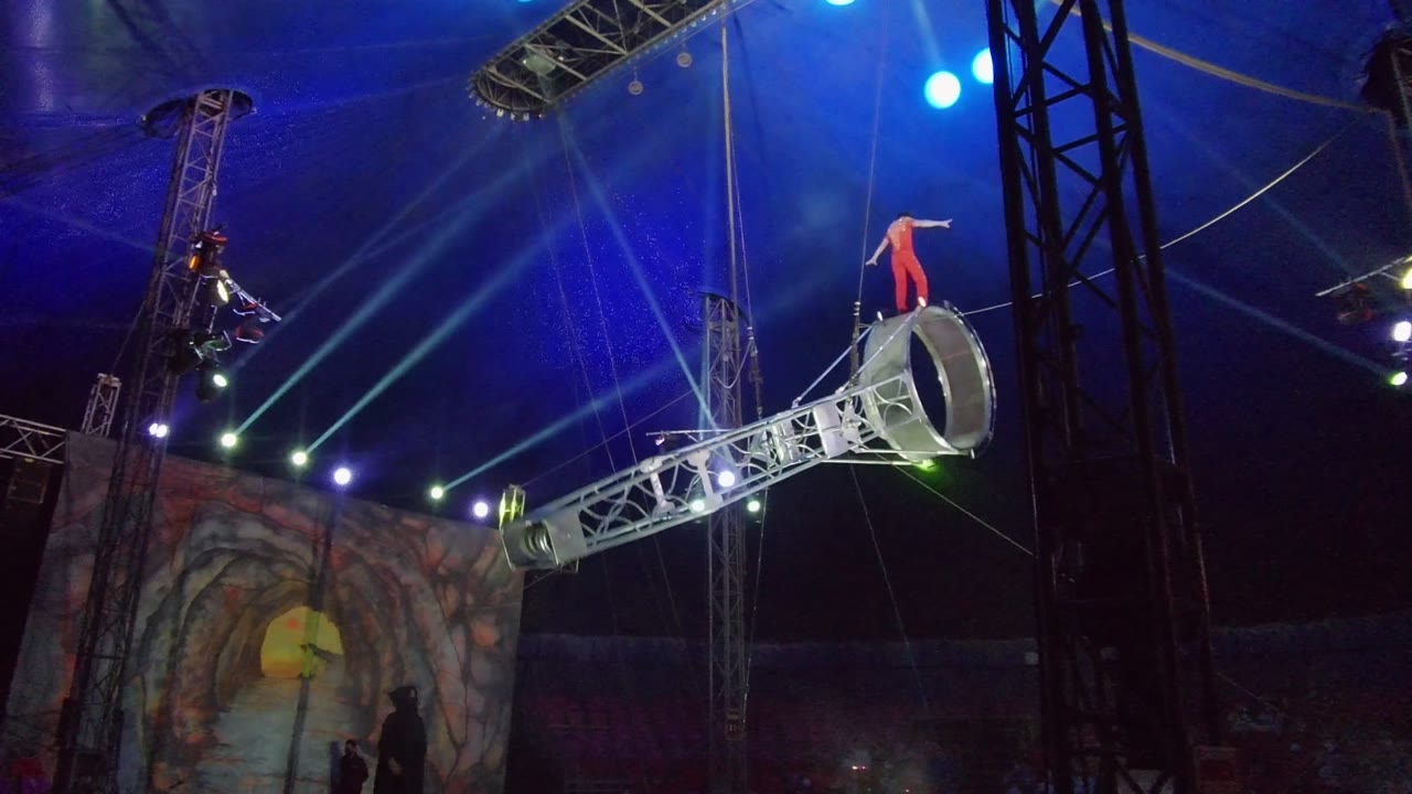 Red-clad acrobat standing on the circular end of a raised truss apparatus.