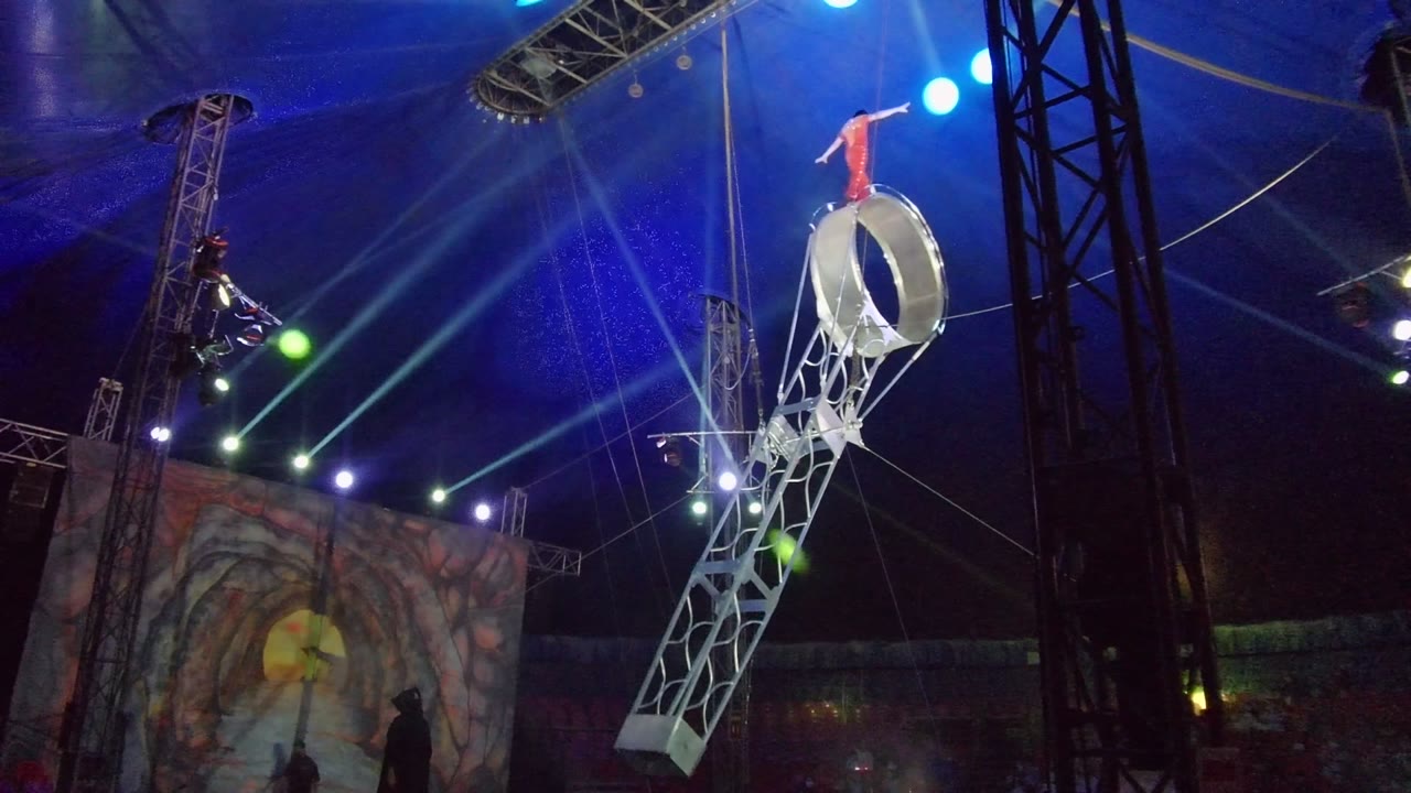 The apparatus has rotated nearly upright in San Jose del Cabo, with the acrobat balanced on top and bright blue spotlights and black stage supports behind the act.