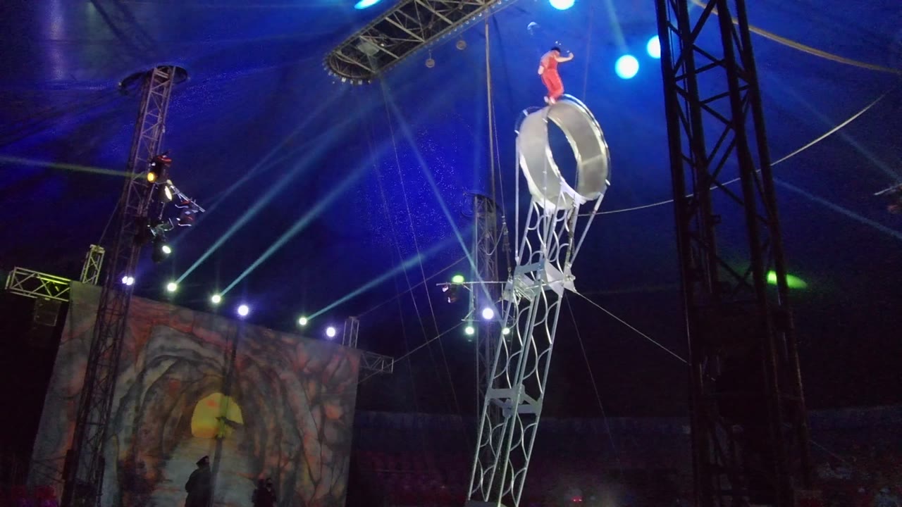 In San Jose Del Cabo, Mexico, a performer in red stands high on a silver aerial wheel suspended by rigging under blue circus tent lights.