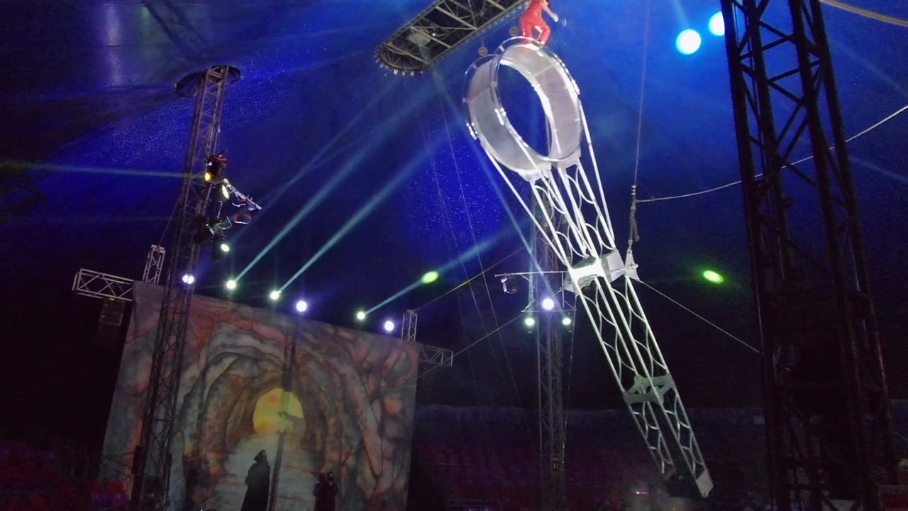 In San Jose del Cabo, Baja California Sur, a performer in red stands near the top of a suspended circular aerial ring while blue and white stage beams cross the tent ceiling.