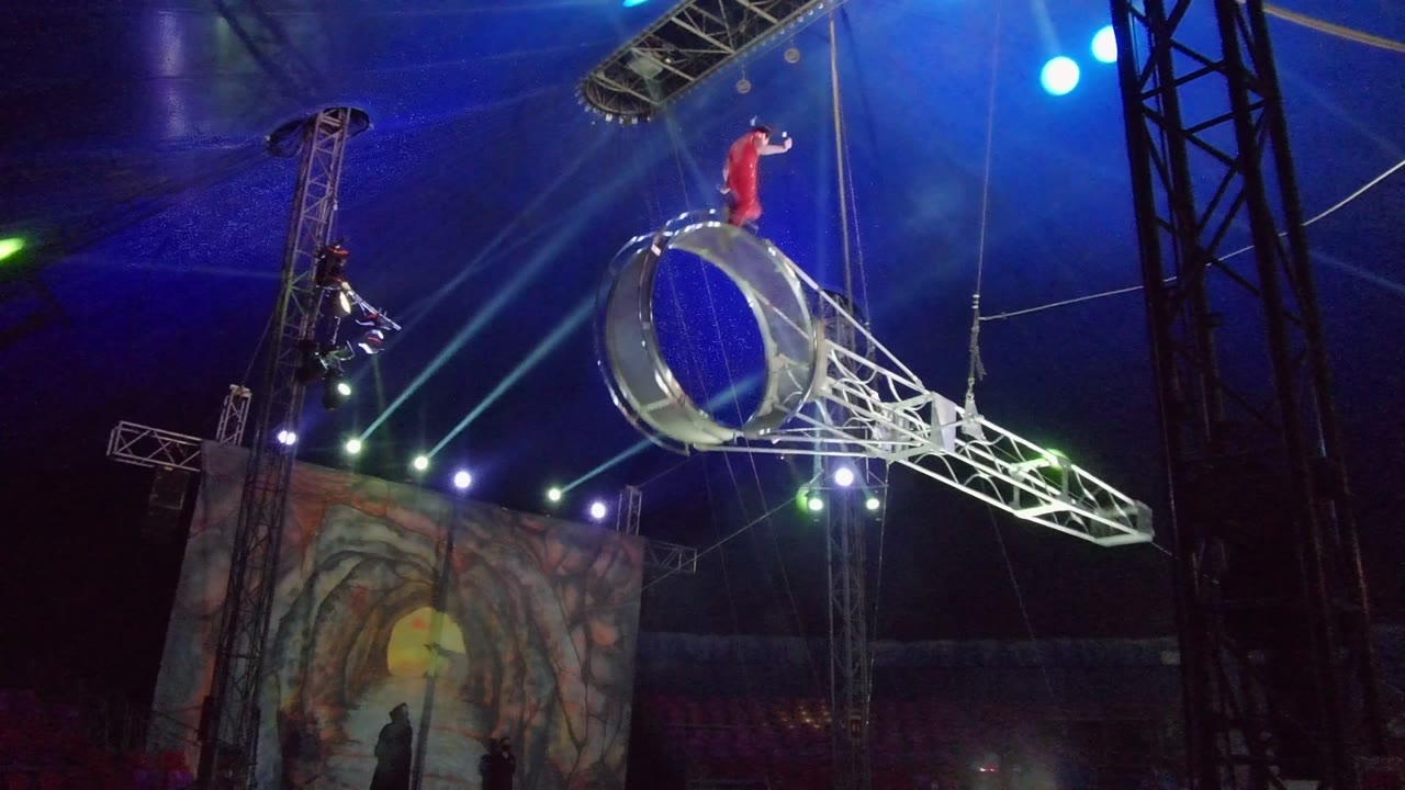 Aerial performer stands on the rotating ring apparatus inside a tent.