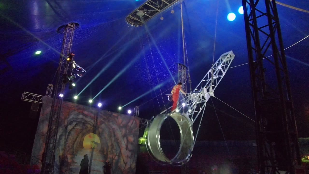 In San Jose del Cabo, Baja California Sur, the aerial ring tilts with the performer in red positioned above it, while multiple stage beams rake across the dark tent interior.