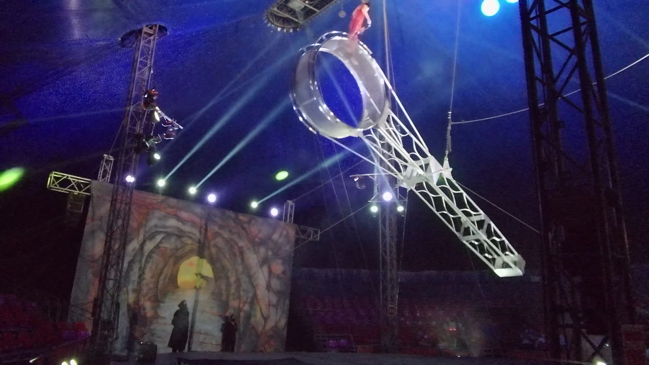 In San Jose del Cabo, a red-clad aerialist appears above a large silver ring on a white truss while blue spotlights cross the circus tent interior.