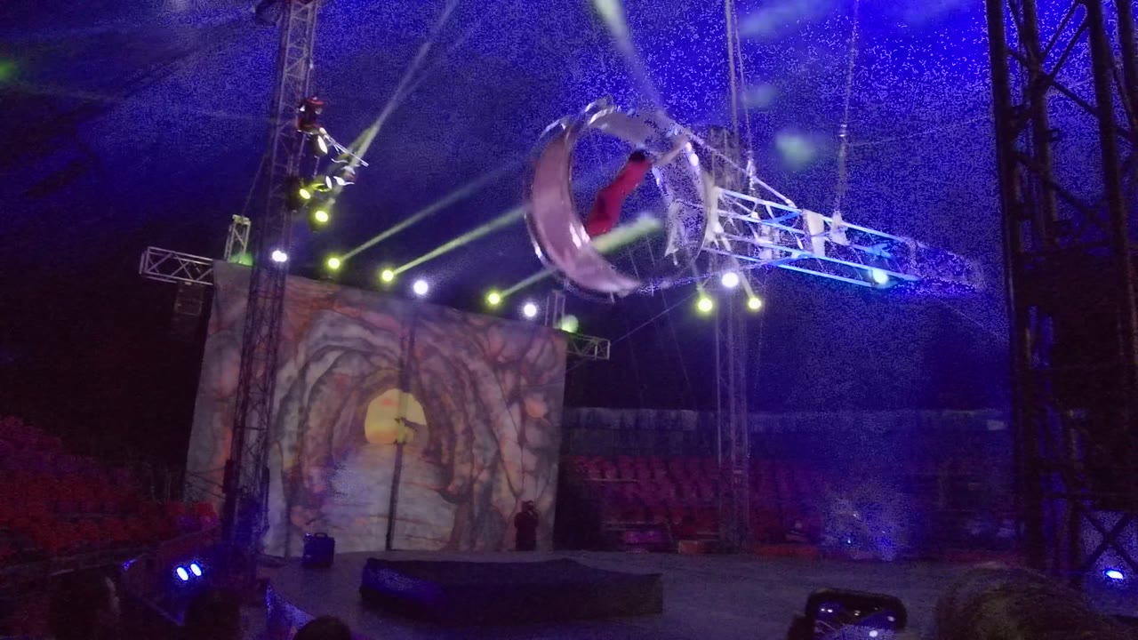 In San Jose del Cabo, a red-clad aerial performer appears inside a tilted circular rig above a stage with blue wash lighting, green beams, red seats, and a scenic screen behind.