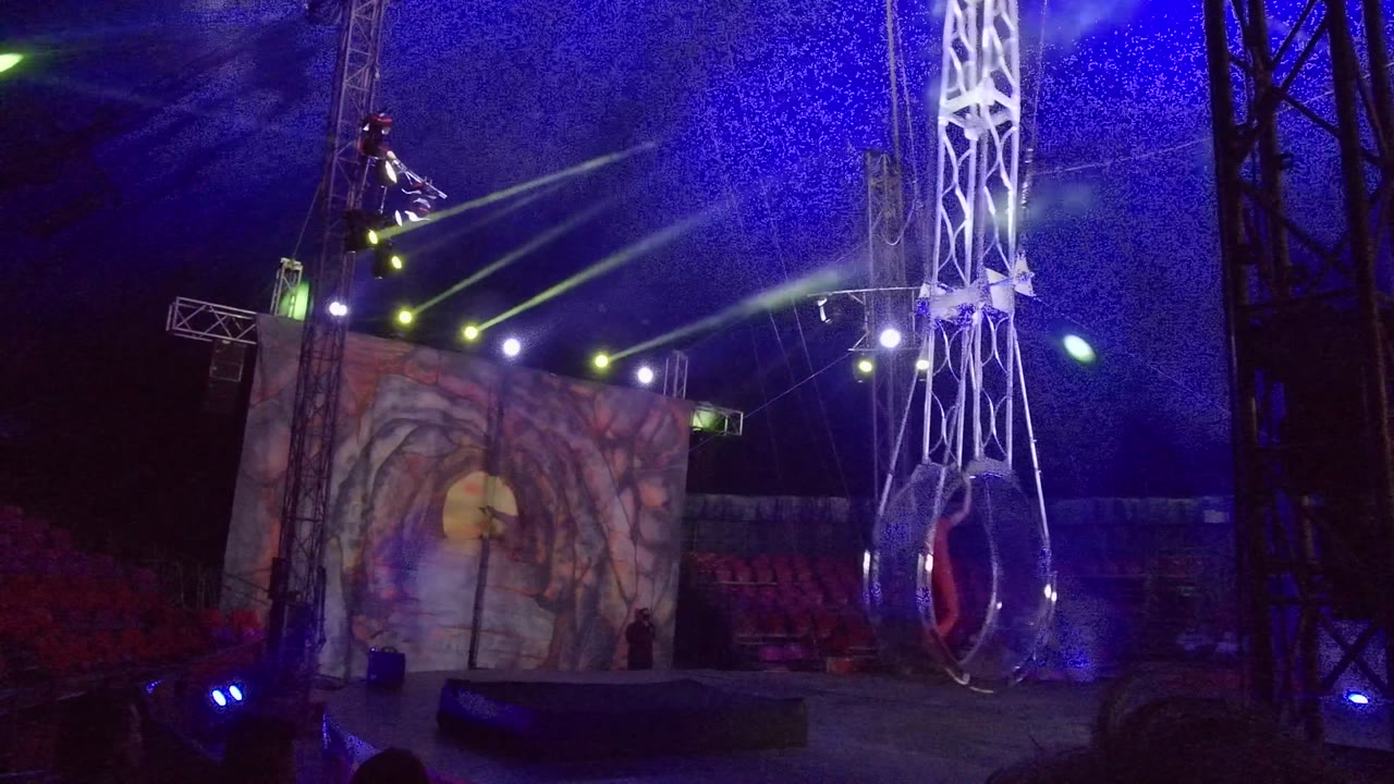 Circular aerial rig hangs near center frame beside tall stage truss.