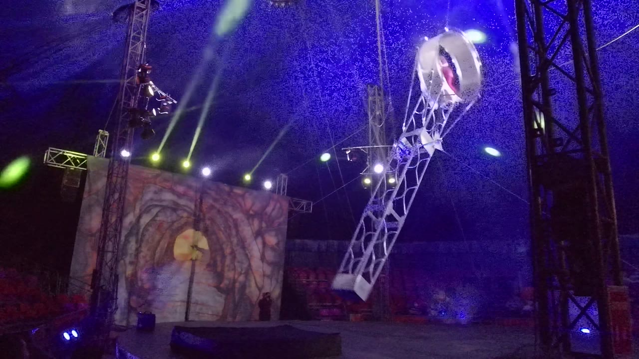 In San Jose del Cabo, Mexico, an aerial performer is suspended in a circular ring on a white metal arm above a stage with blue tent lighting, green spotlights, a painted backdrop, and red seats.