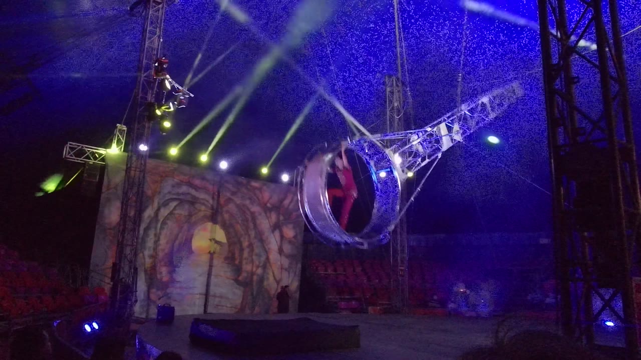 The performer in the circular ring is near the right side of the San Jose del Cabo stage view, with the white metal arm stretching diagonally across purple-blue light and a painted screen behind.