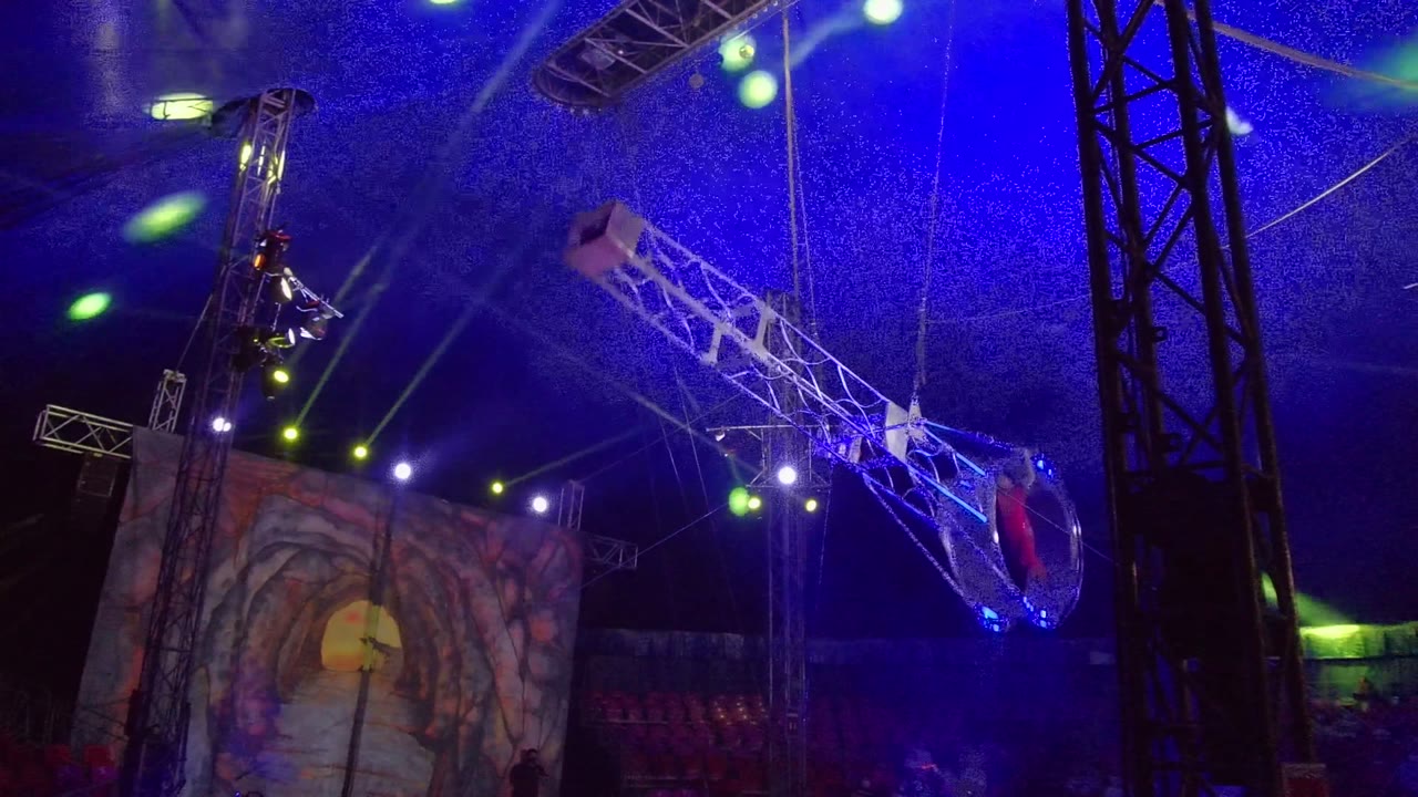 At 0:00 in San Jose del Cabo, an aerial performer in red appears low in a circular hoop attached to a white truss, with blue ceiling wash, green spotlights, black rigging, and a painted backdrop visible.