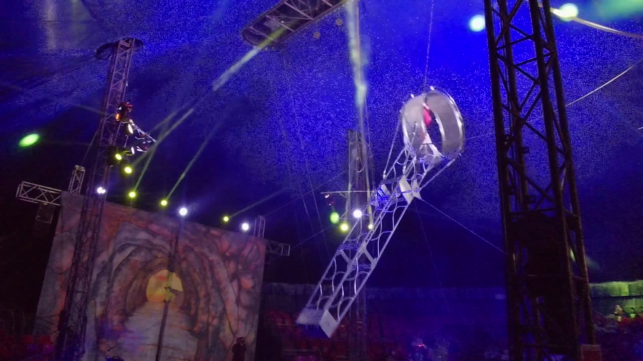 Circular hoop rises near a vertical truss with the performer inside under colored lights.