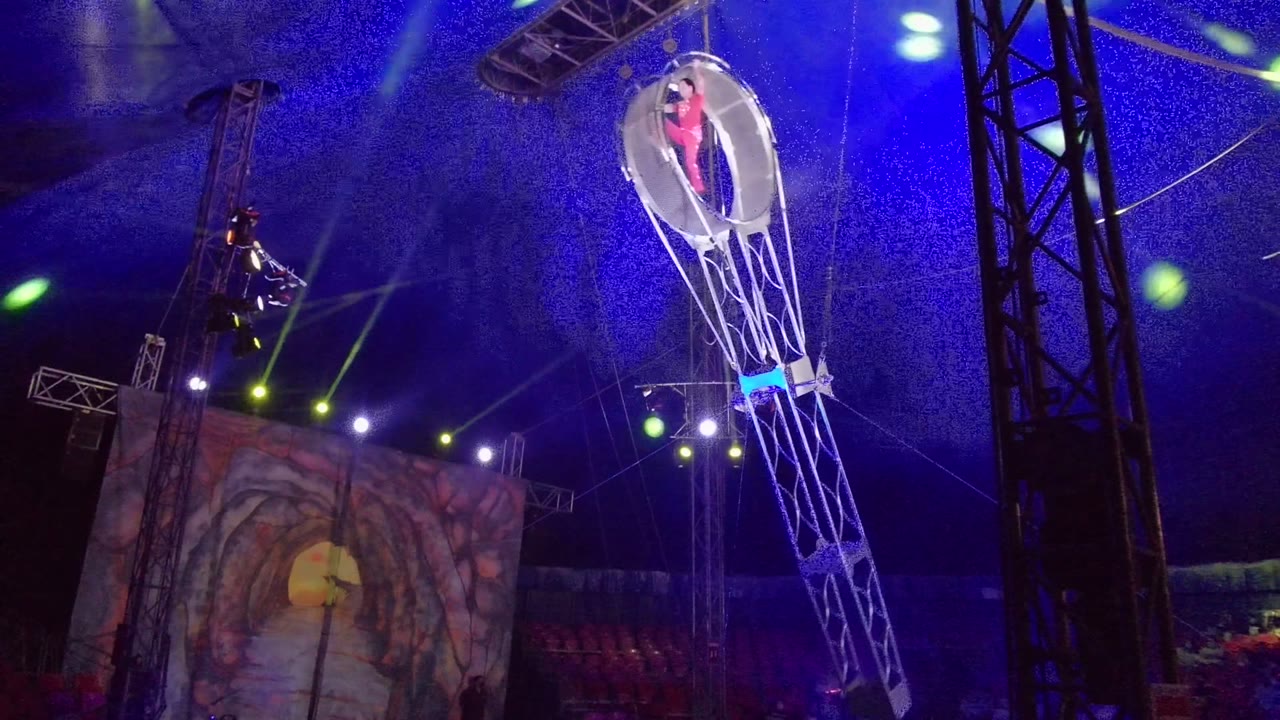 At 0:02 in San Jose del Cabo, the circular hoop reaches the upper right of the frame with the performer standing inside, while blue lighting, green spot beams, black truss columns, and the painted stage backdrop fill the venue.