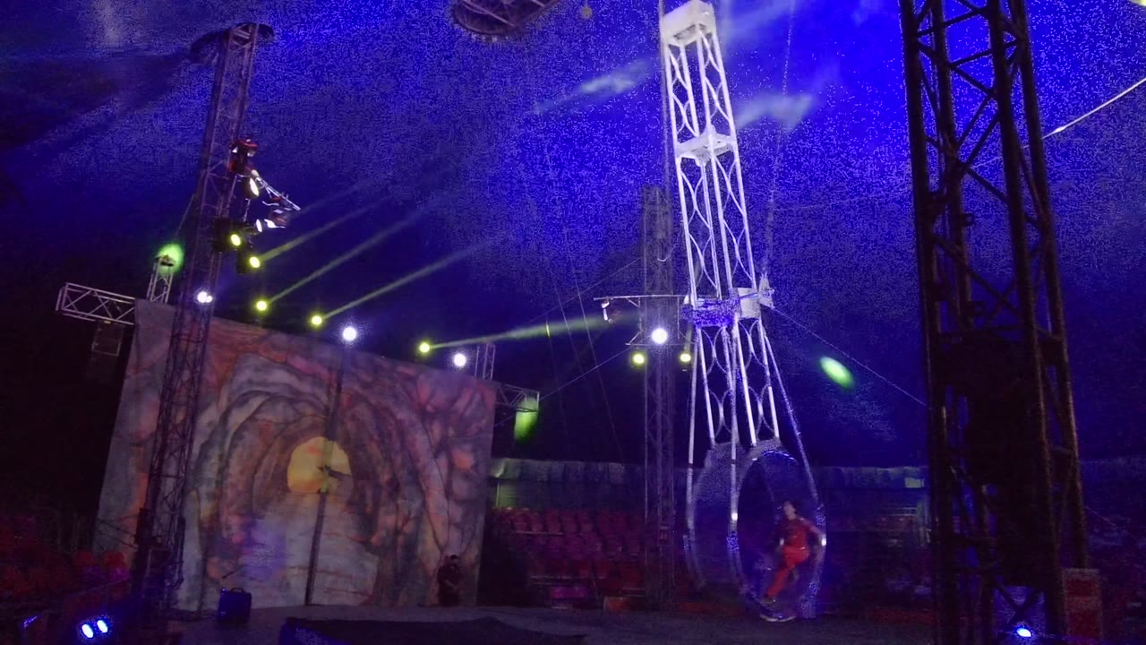 In San Jose Del Cabo, a large suspended circular ring rig stands nearly vertical beside black truss columns, with blue stage light, green beams, red seats, and a tunnel-like screen in the background.