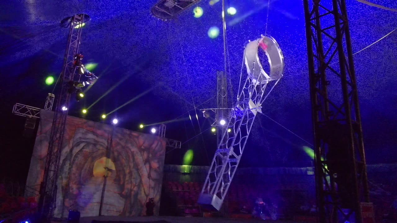 In San Jose Del Cabo, the circular end of the suspended rig is raised high at the right side of frame, with the white frame slanting downward above the stage, blue light, and a performer visible near the upper rigging.