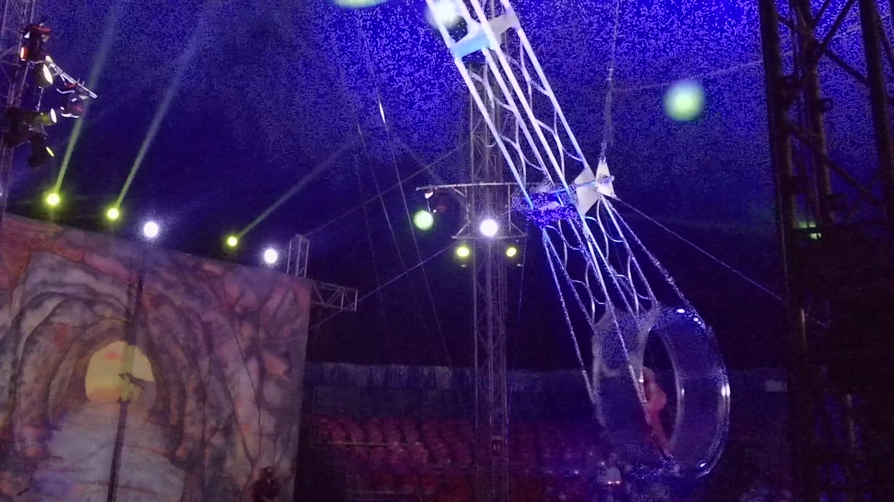 At 0:00 in San Jose Del Cabo, a circular circus cage hangs low on a white mechanical arm beneath blue netting and green stage beams, with part of the performer visible near the cage.