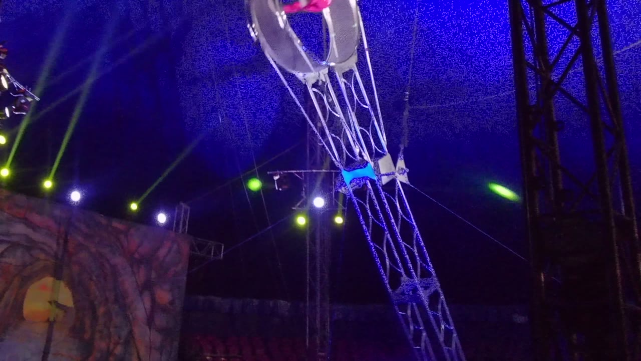 At 0:02 in San Jose Del Cabo, the circular cage is near the top edge of the frame on the mechanical arm, with blue netting overhead, green beams across the stage, and only a partial view of the performer.