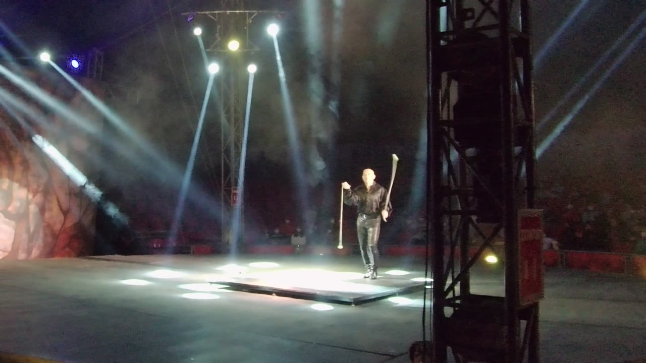 A performer in San Jose Del Cabo stands on a lit stage holding a staff, with white beams crossing through haze and a dark truss at the right edge.