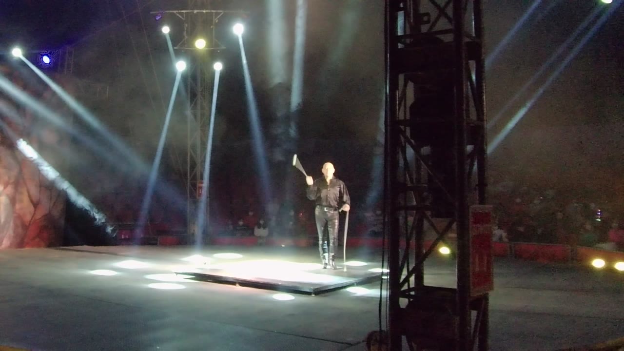 In San Jose del Cabo, a stage performer in dark clothing holds a staff on a lit platform while white spotlights cut through haze above a dark audience area.