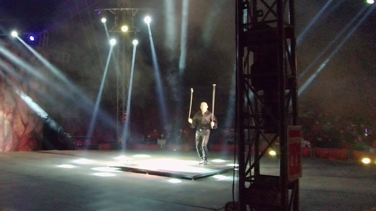 In San Jose del Cabo, the performer appears near the right edge of the platform with the staff upright while bright white beams and haze dominate the dark venue.