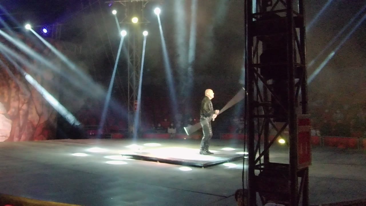 In San Jose del Cabo, Mexico, a solo performer in dark clothing stands on a lit stage holding a large prop while white beams shine through haze behind them.