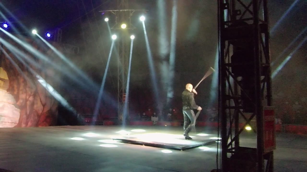 In San Jose del Cabo, Mexico, a dark-clad stage performer raises a large axe on a low platform as white spotlights sweep through smoke behind him.