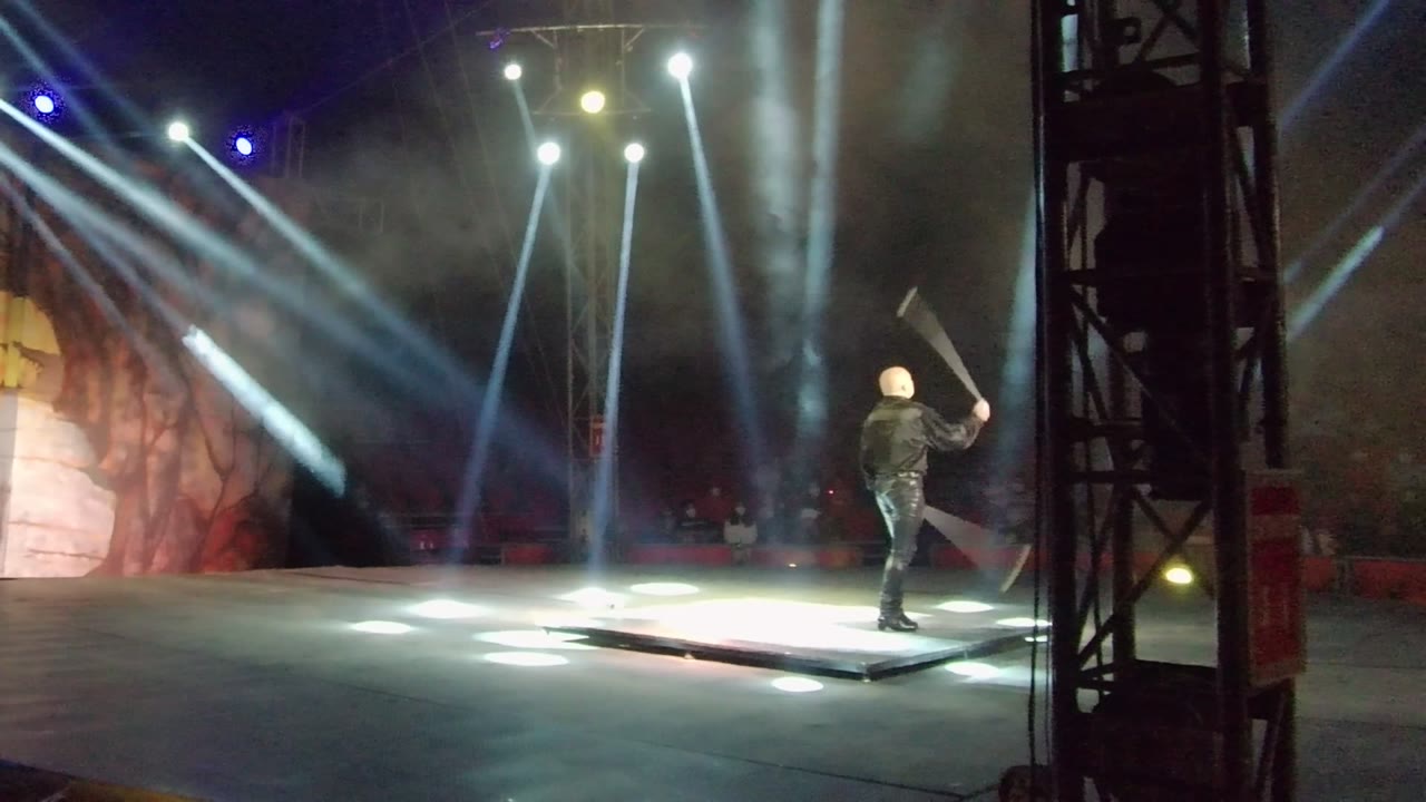 In San Jose del Cabo, Mexico, the performer moves toward the far right edge of the frame while swinging the axe through bright angled spotlights and haze.