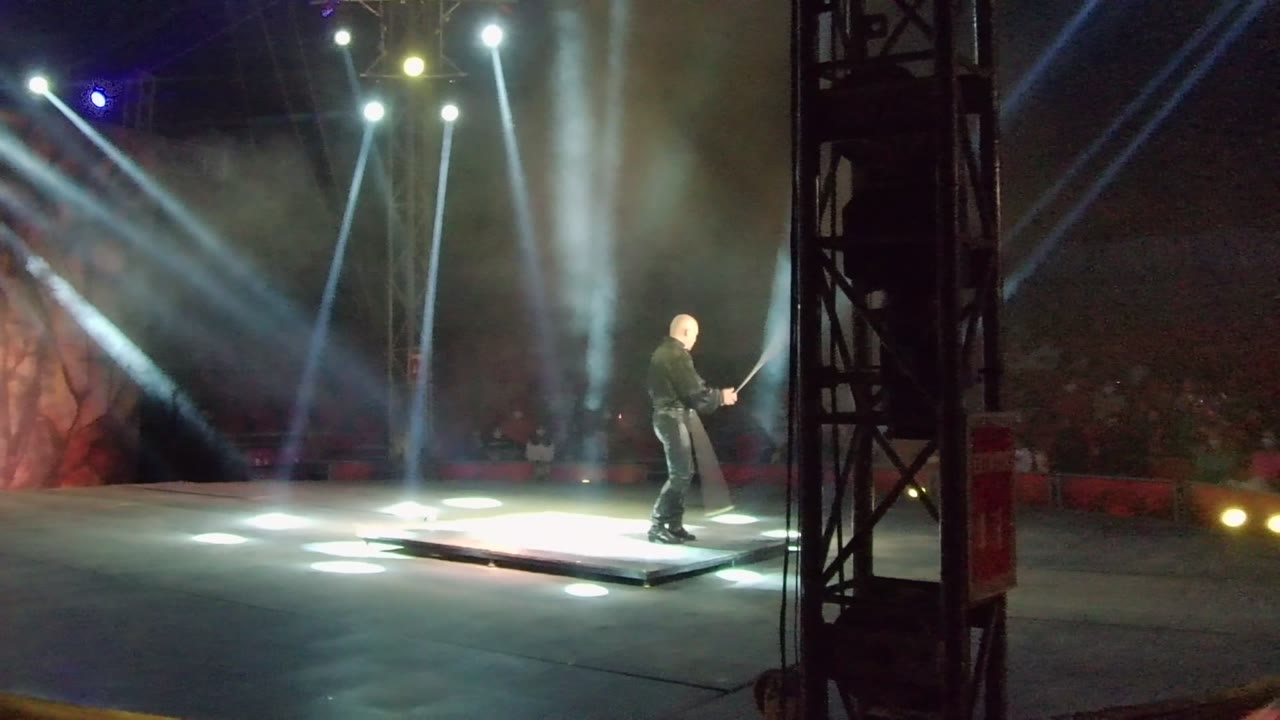 In San Jose Del Cabo, Mexico, a black-clad stage performer stands on a lit platform while swinging a pale flag under tall white beams and haze.