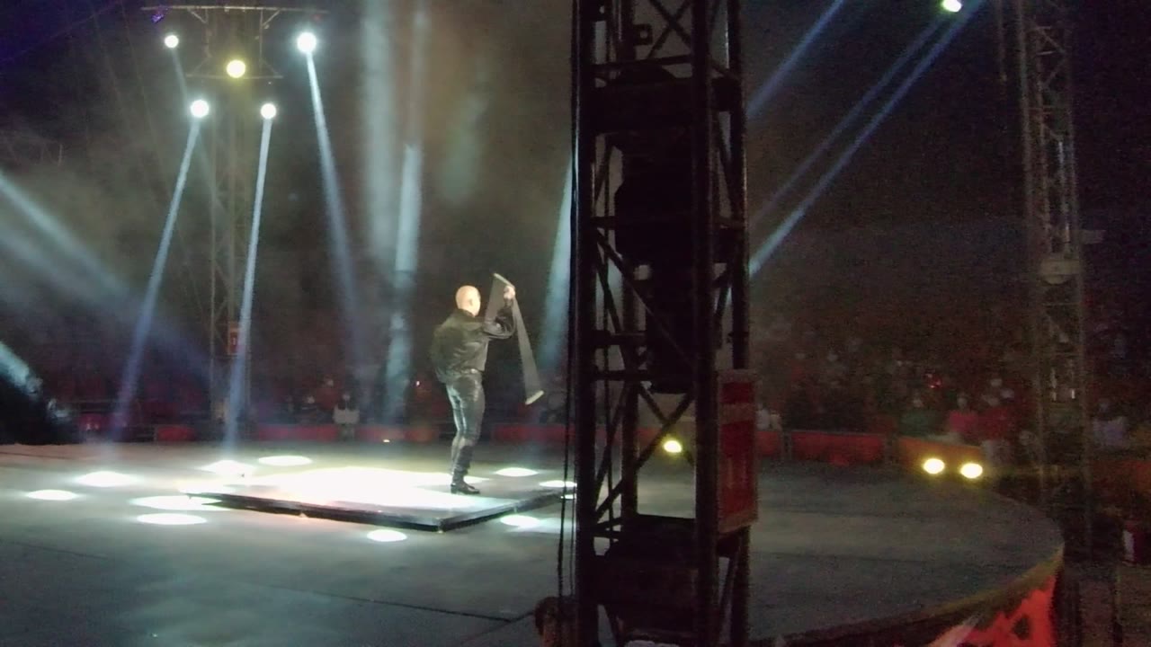 In San Jose Del Cabo, a solo stage performer in dark clothing raises a large prop on a lit platform while white spotlights and haze fill the dark venue.