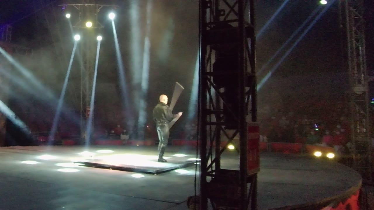 Performer stands on stage with a long prop beside spotlights.